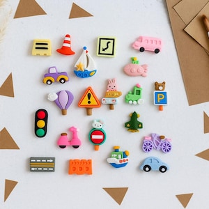 May include: A collection of colorful miniature transportation and road sign-themed decorations. Includes a pink bus, a purple car, a sailboat, a hot air balloon, a traffic light, and a scooter. The decorations are arranged on a white surface.