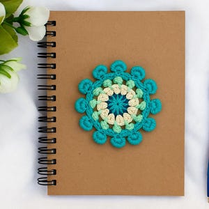Puede incluir: Un cuaderno con espiral marrón con una flor de ganchillo turquesa, crema y verde claro. Un bolígrafo azul descansa junto al cuaderno sobre una superficie blanca, con flores blancas en la esquina superior izquierda. El cuaderno es un artículo artesanal.