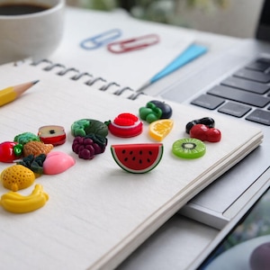 May include: A collection of small, colorful fruit-shaped charms scattered on a spiral-bound notebook. The charms include miniature representations of watermelon, kiwi, grapes, bananas, apples, and other assorted fruits in vibrant plastic finishes.