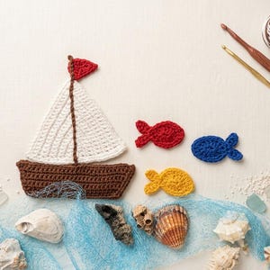 May include: A nautical-themed crochet arrangement featuring a brown boat with a white sail and red flag, alongside red, blue, and yellow fish. Seashells, sea glass, and a blue net are arranged on a white surface, with crochet hooks and yarn in the background.