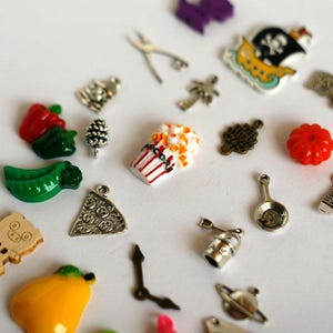 Puede incluir: Una colección de pequeños abalorios y colgantes de colores. Incluye una caja de palomitas de maíz, una porción de pizza, un pimiento rojo, una calabaza, un cerdo y otros objetos en miniatura. Los abalorios están dispersos sobre una superficie blanca.