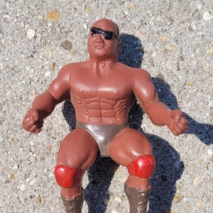 Könnte beinhalten: Eine braune Plastik-Actionfigur eines Wrestlers mit schwarzer Sonnenbrille, braunen Stiefeln und roten Knieschonern. Die Figur sitzt mit gespreizten Beinen.
