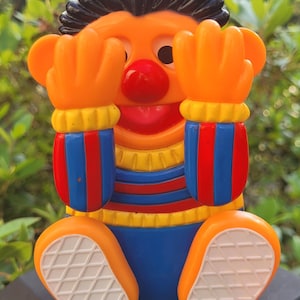 May include: A colorful plastic toy figure with orange skin, black hair, and a red nose. The figure has orange hands covering its eyes, blue and red striped sleeves, and blue pants. The figure's feet are orange with white soles. The toy is a children's collectible.