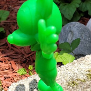 Op de afbeelding: Een groen plastic beeldje van Mickey Mouse dat op een met mos bedekte rots staat. Mickey draagt een groen shirt en broek en heeft zijn rechterhand opgeheven in een groet.