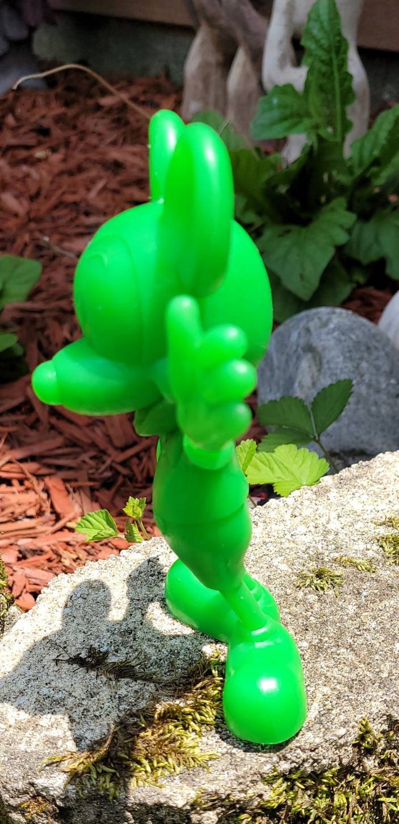 Op de afbeelding: Een groen plastic beeldje van Mickey Mouse dat op een met mos bedekte rots staat. Mickey draagt een groen shirt en broek en heeft zijn rechterhand opgeheven in een groet.