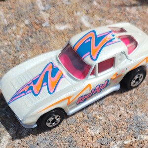 May include: White toy car with a pink, blue, and orange swirl design. The car has a clear windshield and the words "Kool-Aid" on the side.