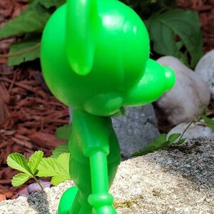 Op de afbeelding: Een felgroen plastic beeldje van een cartoonfiguur. Het figuur staat met zijn armen langs zijn zijde en zijn hoofd gekanteld naar de zijkant. Het beeldje staat op een grijs, getextureerd oppervlak.