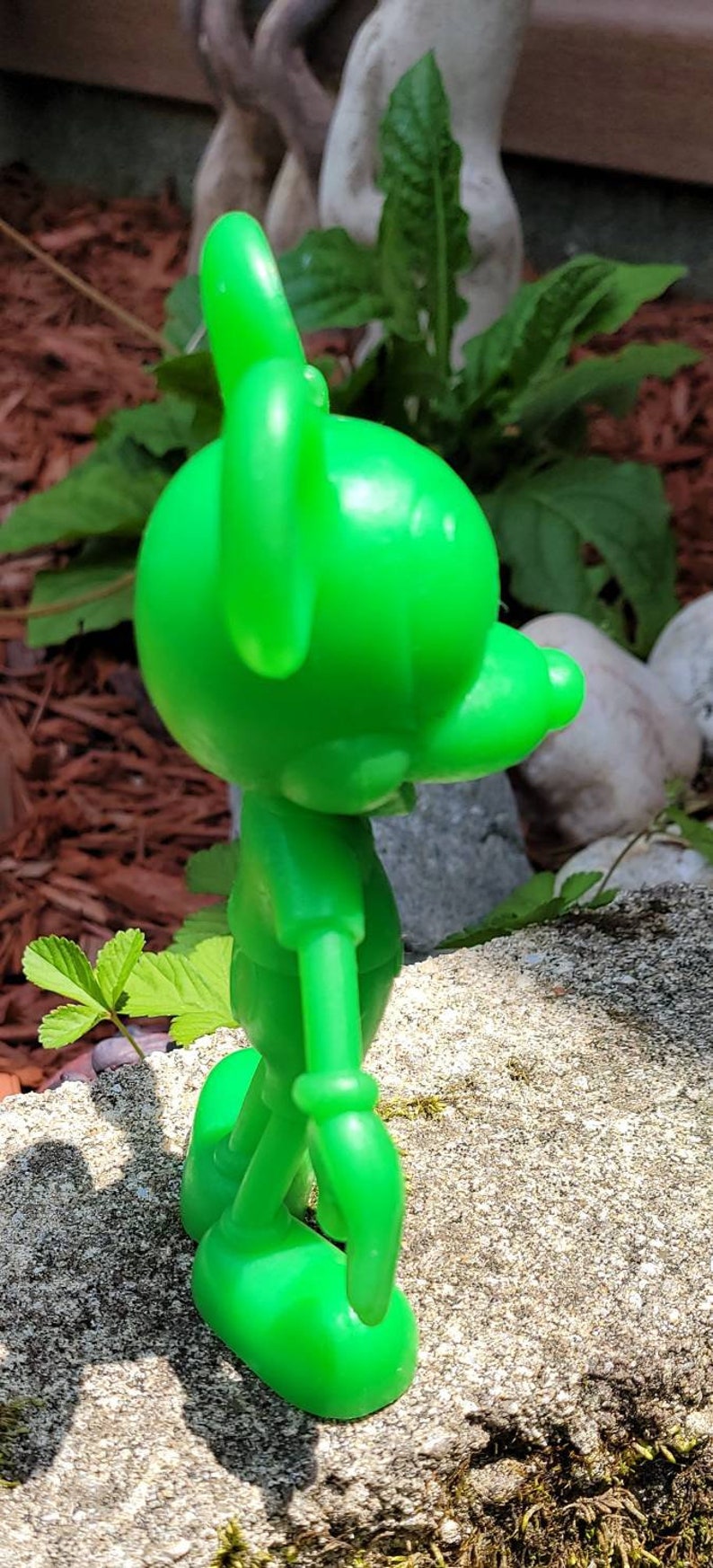 Op de afbeelding: Een felgroen plastic beeldje van een cartoonfiguur. Het figuur staat met zijn armen langs zijn zijde en zijn hoofd gekanteld naar de zijkant. Het beeldje staat op een grijs, getextureerd oppervlak.