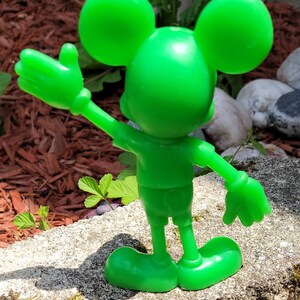 Op de afbeelding: Een felgroen plastic beeldje van Mickey Mouse met zijn arm omhoog in een zwaai. Het beeldje staat op een grijs rotsoppervlak.