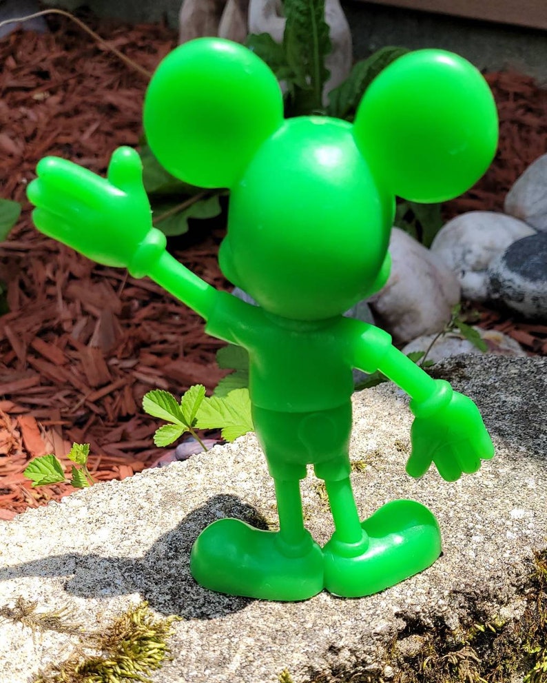 Op de afbeelding: Een felgroen plastic beeldje van Mickey Mouse met zijn arm omhoog in een zwaai. Het beeldje staat op een grijs rotsoppervlak.