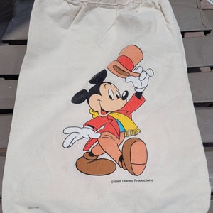 Puede incluir: Una bolsa de lona blanca con cord&oacute;n de ajuste con una ilustraci&oacute;n de Mickey Mouse. Mickey lleva una chaqueta roja, una bufanda amarilla y zapatos marrones. Est&aacute; sosteniendo un sombrero marr&oacute;n y rojo en su mano derecha. La bolsa tiene el texto "&copy; Walt Disney Productions" impreso en la parte inferior. La bolsa tambi&eacute;n tiene el texto "Made in USA" impreso en la parte inferior.