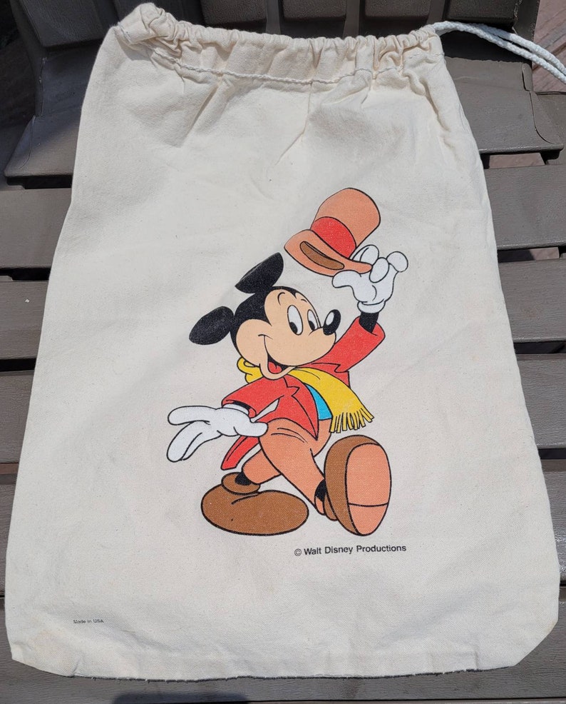 Puede incluir: Una bolsa de lona blanca con cord&oacute;n de ajuste con una ilustraci&oacute;n de Mickey Mouse. Mickey lleva una chaqueta roja, una bufanda amarilla y zapatos marrones. Est&aacute; sosteniendo un sombrero marr&oacute;n y rojo en su mano derecha. La bolsa tiene el texto "&copy; Walt Disney Productions" impreso en la parte inferior. La bolsa tambi&eacute;n tiene el texto "Made in USA" impreso en la parte inferior.
