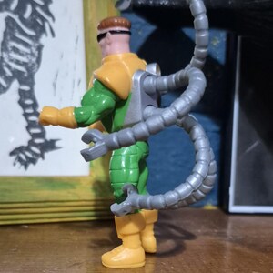 May include: A Doctor Octopus action figure. The figure is wearing a green and yellow outfit with grey mechanical arms extending from its back. The figure has brown hair and sunglasses. The figure is standing on a wooden surface.