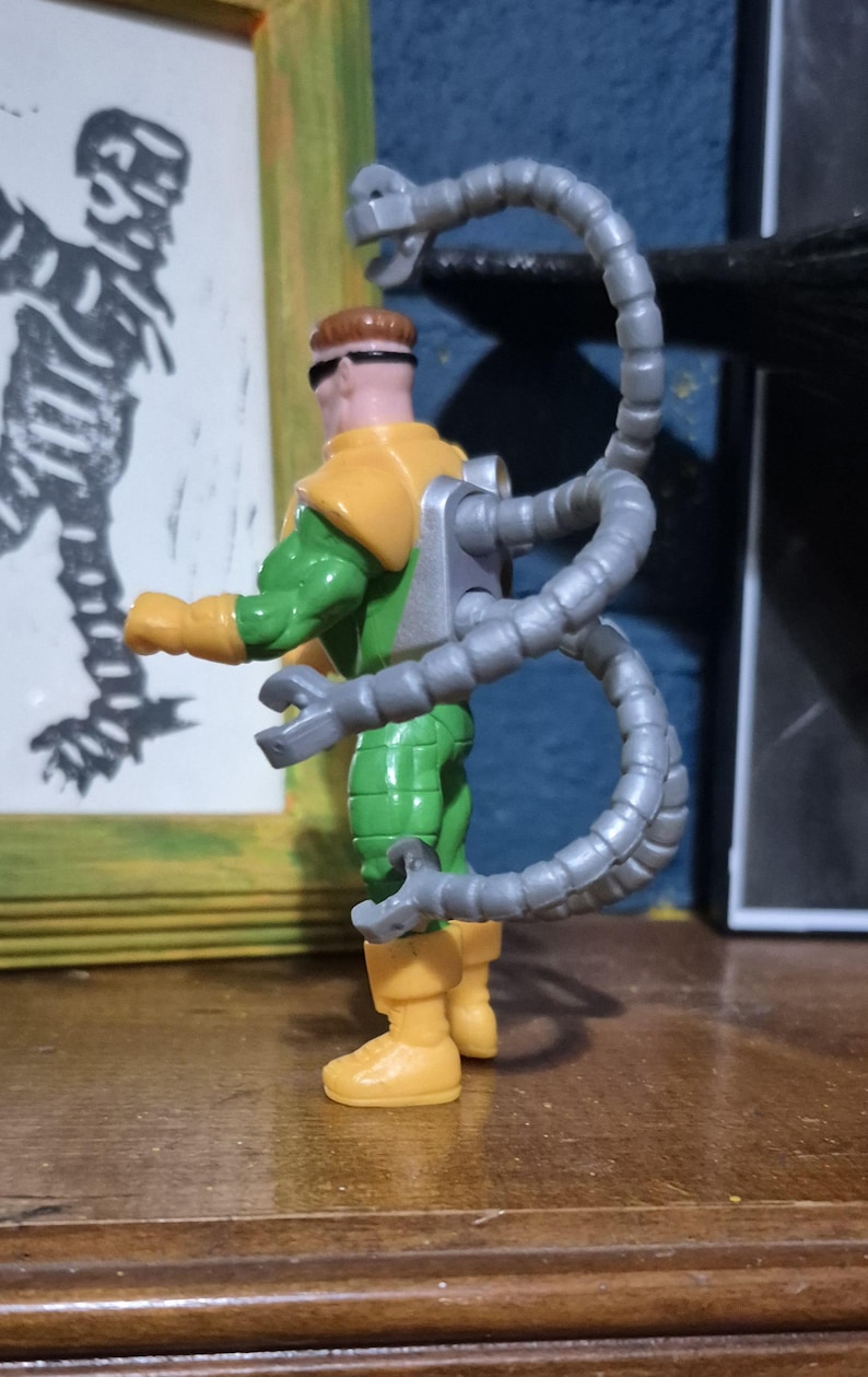 May include: A Doctor Octopus action figure. The figure is wearing a green and yellow outfit with grey mechanical arms extending from its back. The figure has brown hair and sunglasses. The figure is standing on a wooden surface.