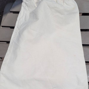 Puede incluir: Una bolsa de algod&oacute;n blanco con un cord&oacute;n de ajuste blanco. La bolsa est&aacute; tendida sobre una superficie marr&oacute;n.