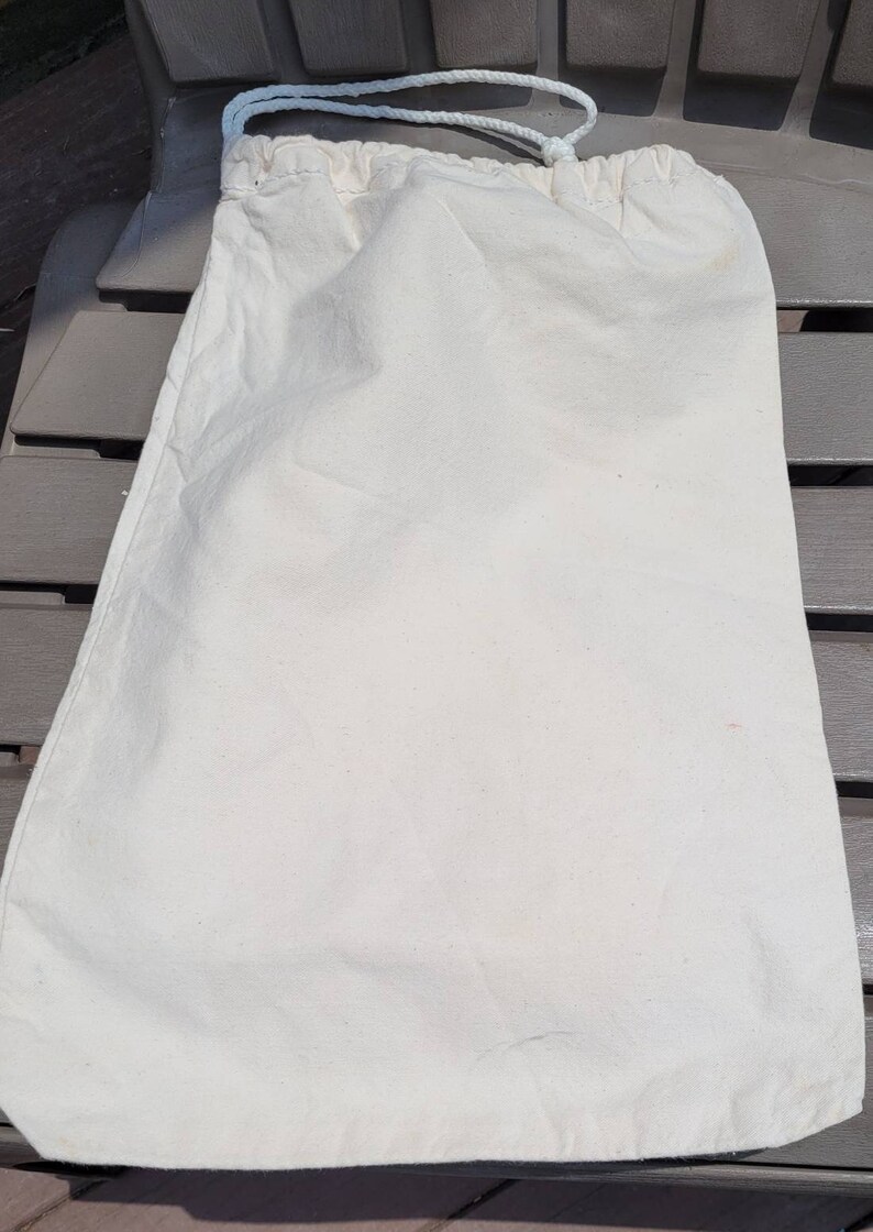 Puede incluir: Una bolsa de algod&oacute;n blanco con un cord&oacute;n de ajuste blanco. La bolsa est&aacute; tendida sobre una superficie marr&oacute;n.
