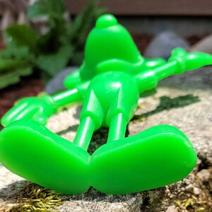 Op de afbeelding: Een felgroen plastic speelgoedfiguur van een cartoonfiguur. De figuur ligt op zijn rug met zijn armen uitgestrekt en zijn benen gebogen in de knie&euml;n. De figuur heeft een groot hoofd met een rond gezicht en een kleine neus. De voeten van de figuur zijn zichtbaar op de voorgrond.