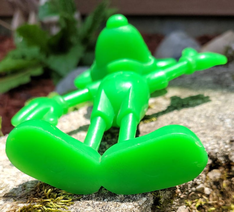Op de afbeelding: Een felgroen plastic speelgoedfiguur van een cartoonfiguur. De figuur ligt op zijn rug met zijn armen uitgestrekt en zijn benen gebogen in de knie&euml;n. De figuur heeft een groot hoofd met een rond gezicht en een kleine neus. De voeten van de figuur zijn zichtbaar op de voorgrond.