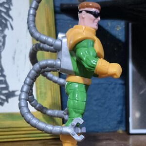 May include: A Dr. Octopus action figure. The figure is wearing a green and yellow suit with silver mechanical arms. The figure has brown hair and sunglasses. The figure is standing on a wooden surface.