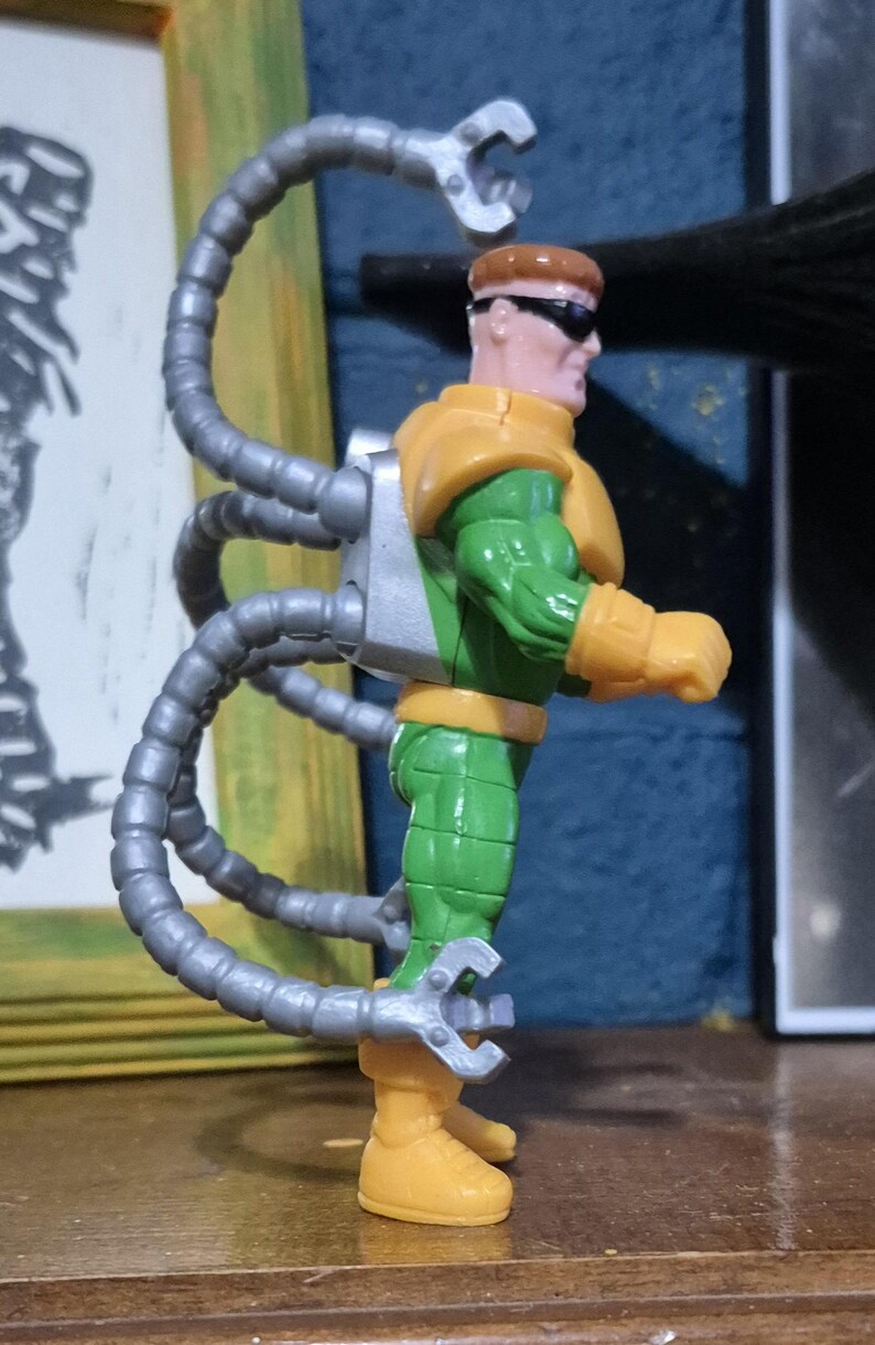 May include: A Dr. Octopus action figure. The figure is wearing a green and yellow suit with silver mechanical arms. The figure has brown hair and sunglasses. The figure is standing on a wooden surface.