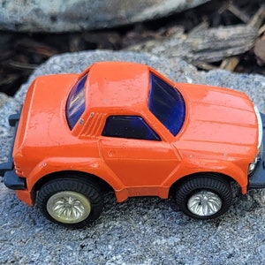 May include: Orange toy car with black accents and silver wheels. The car has a blue tinted windscreen and a detailed interior.