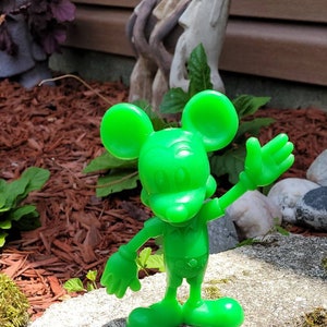 Op de afbeelding: Een felgroen plastic beeldje van Mickey Mouse met een vriendelijke uitdrukking, zwaaiend met zijn rechterhand. Het beeldje staat op een grijze steen bedekt met mos.