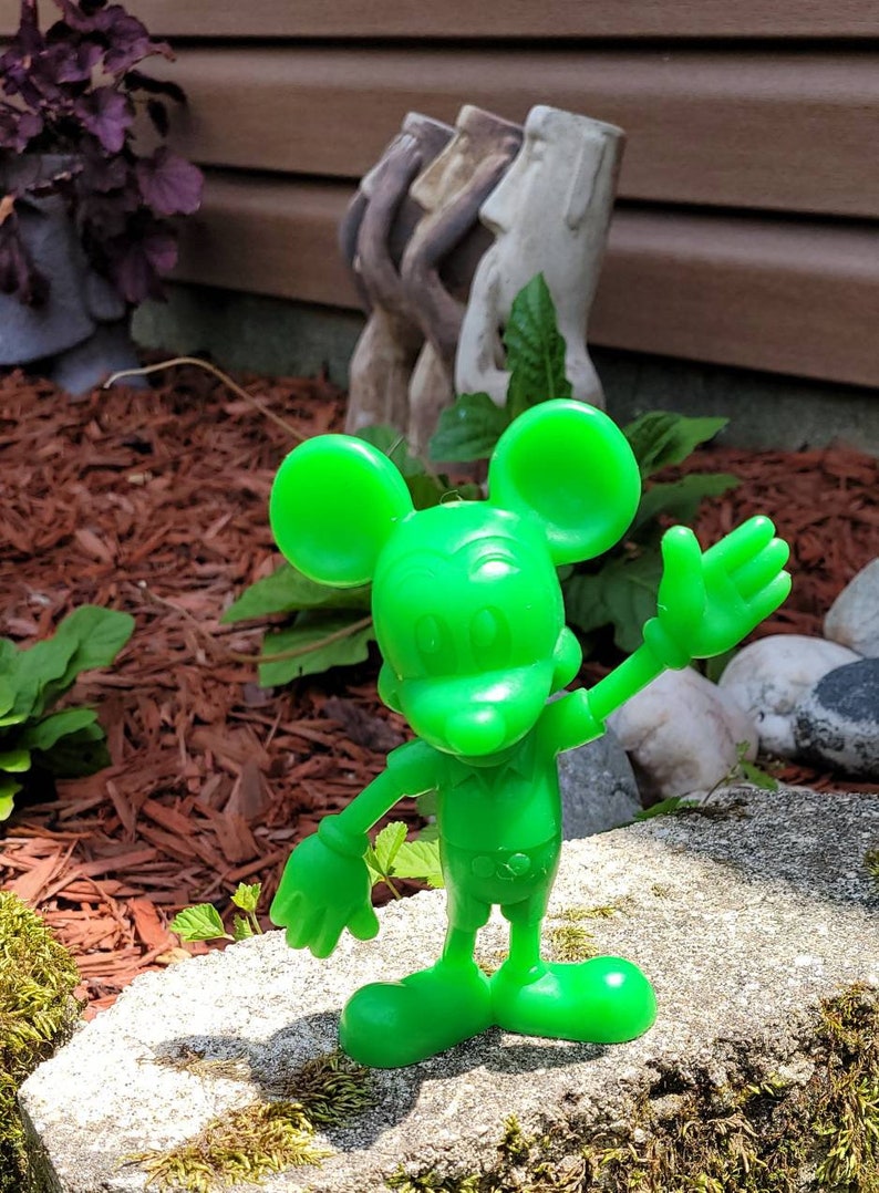 Op de afbeelding: Een felgroen plastic beeldje van Mickey Mouse met een vriendelijke uitdrukking, zwaaiend met zijn rechterhand. Het beeldje staat op een grijze steen bedekt met mos.