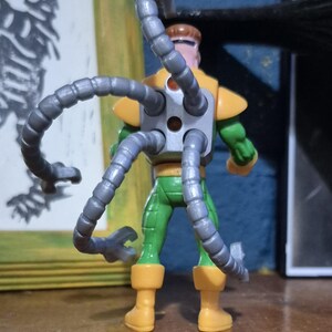 May include: A Dr. Octopus action figure, seen from behind, with mechanical tentacles. The figure is mainly green, with yellow details on the shoulders and boots. The articulated gray tentacles are a key feature of the toy, enhancing its dynamic look.