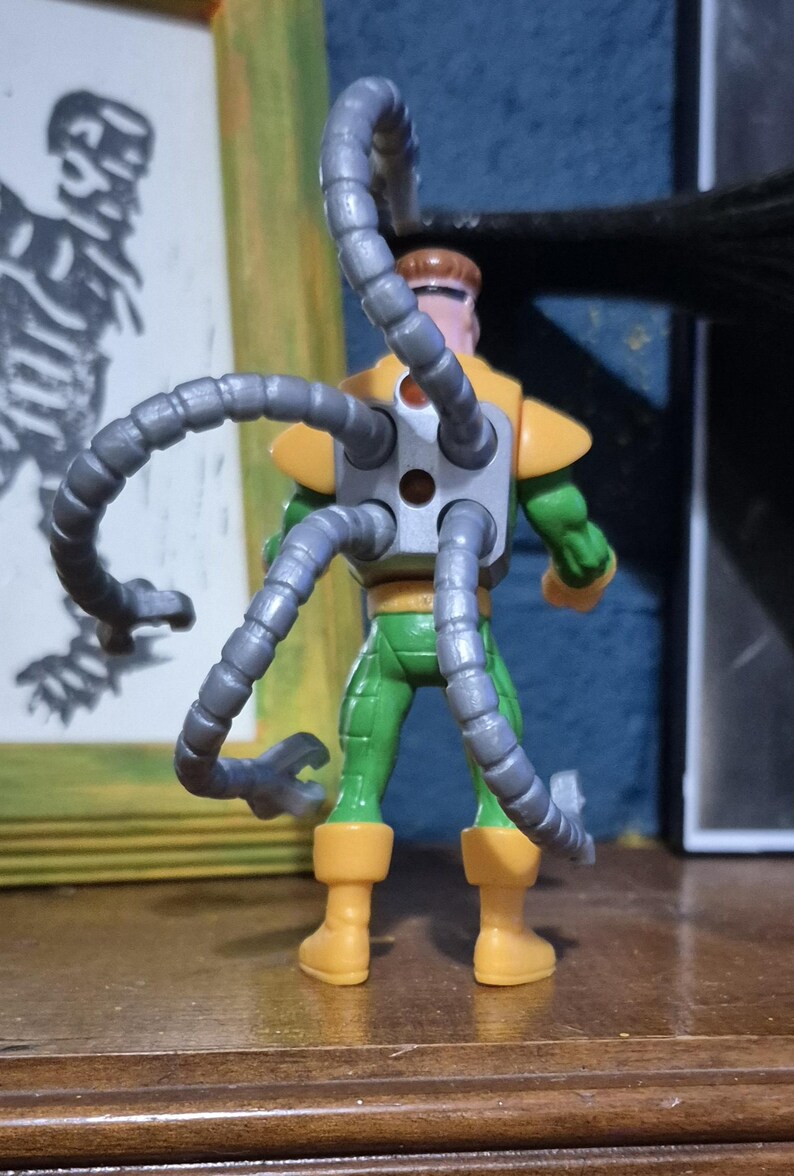 May include: A Dr. Octopus action figure, seen from behind, with mechanical tentacles. The figure is mainly green, with yellow details on the shoulders and boots. The articulated gray tentacles are a key feature of the toy, enhancing its dynamic look.