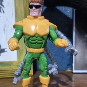 May include: A Dr. Octopus action figure. The figure is wearing a yellow and green outfit with sunglasses. The figure has four mechanical arms attached to its back. The figure is standing on a wooden surface.