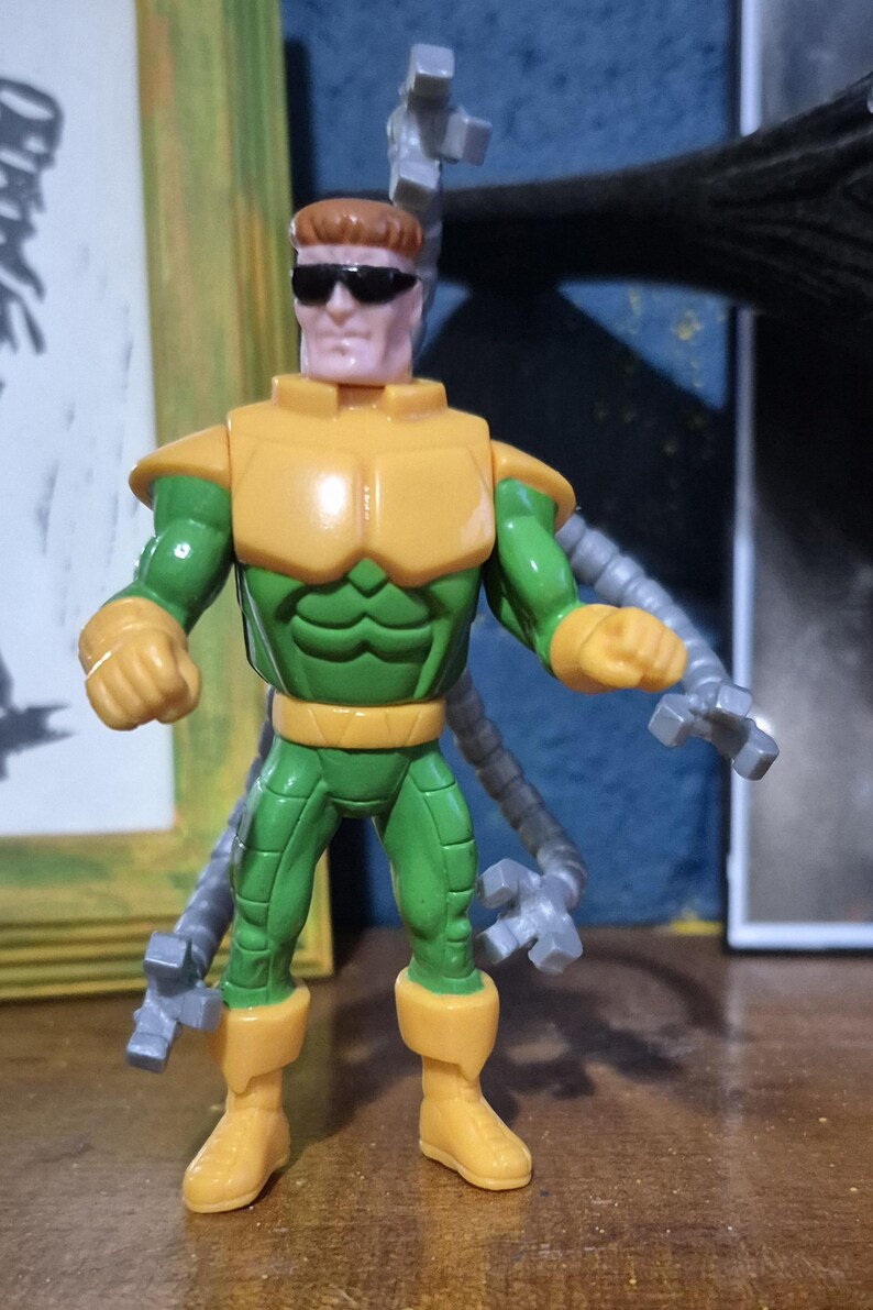 May include: A Dr. Octopus action figure. The figure is wearing a yellow and green outfit with sunglasses. The figure has four mechanical arms attached to its back. The figure is standing on a wooden surface.