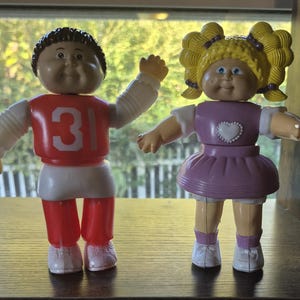 Pode incluir: Duas bonecas Cabbage Patch Kids vintage. Uma boneca está vestindo uma camisa vermelha com o número 31, mangas brancas e calças vermelhas. A outra boneca está vestindo um vestido roxo com um coração branco e cabelo amarelo.