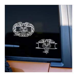 May include: Two white vinyl decals on a vehicle window. One is an oval design with a cross, wings, and a snake. The other is a similar design, but rectangular. Both have the text "MILVEC".