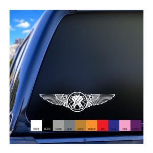May include: White vinyl decal featuring a medical symbol with wings, designed for application on a vehicle window. The emblem includes a star of life symbol at the center, with a pair of crossed tools. The decal is shown against a dark background with color swatches.
