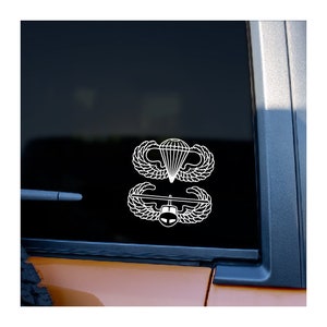 May include: White vinyl decal featuring two military insignia: a parachute with wings and a pilot's badge with wings. The decals are displayed on a vehicle window, offering a clean, contrasting look against the dark glass.