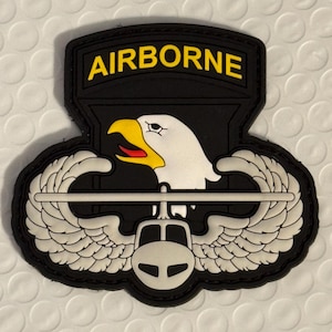 May include: A black patch with the word "AIRBORNE" in yellow block letters. Below the text is a white eagle head with a yellow beak and red tongue. The patch also features white wings and a helicopter.