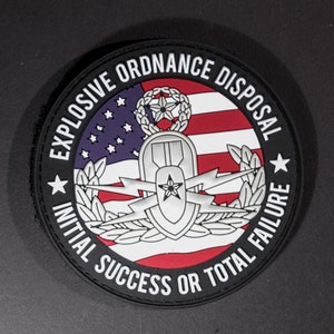 May include: A black and white patch with the text "Explosive Ordnance Disposal Initial Success or Total Failure" around the edge. The center of the patch features a silver EOD badge with a star and lightning bolts, set against a red, white, and blue American flag.