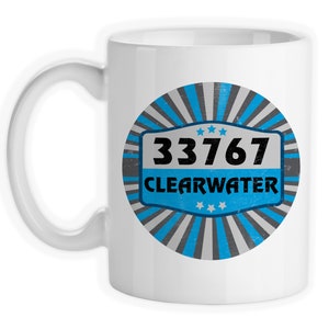 May include: White ceramic coffee mug with a blue and gray circular design. The design features the text "33767 CLEARWATER" with five stars above and below the text.
