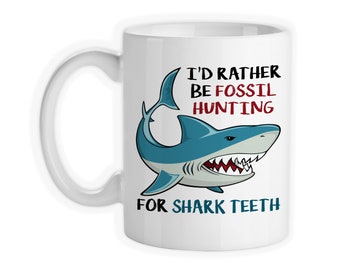 May include: White ceramic coffee mug with a blue shark illustration and the text "I'd rather be fossil hunting for shark teeth".