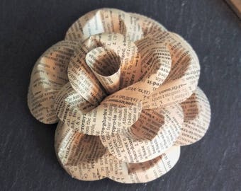 May include: A paper flower crafted from pages of a book, with the text visible. The flower is a light brown color and has a delicate, layered design.