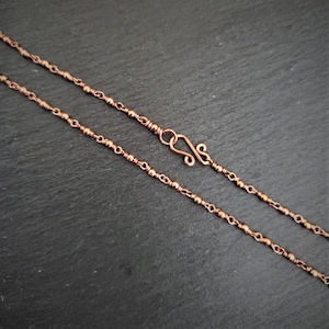 May include: A copper-colored necklace with a delicate chain design. The necklace features small, round beads interspersed along the chain, with a decorative hook clasp. The jewelry is displayed against a dark background.