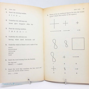 May include: Open book with "TEST ONE" at the top of the pages. The left page has numbered questions, including fill-in-the-blank and odd-one-out problems. The right page has a similar test with geometric shapes. The book is held open by a metal stand.