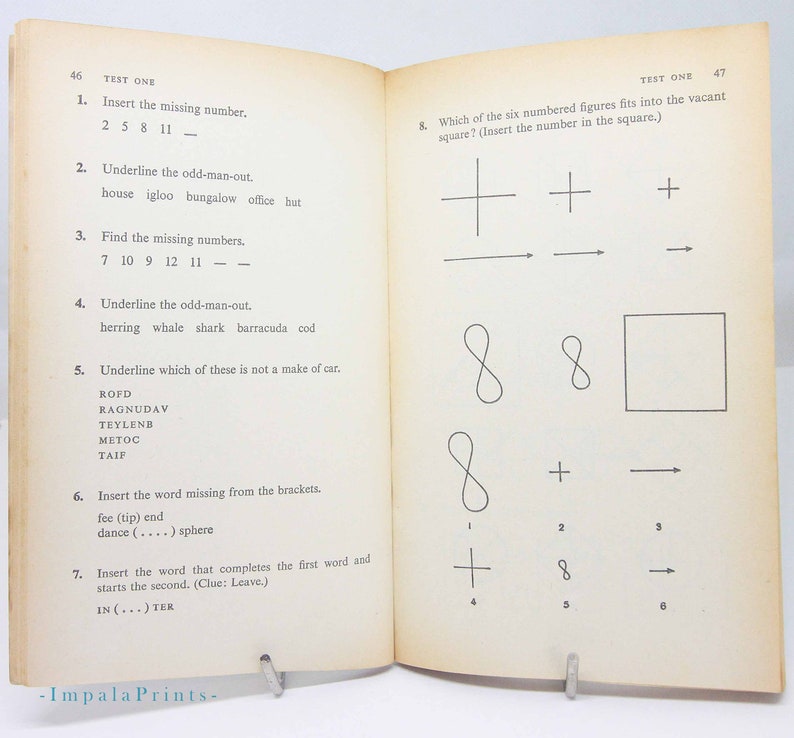 May include: Open book with "TEST ONE" at the top of the pages. The left page has numbered questions, including fill-in-the-blank and odd-one-out problems. The right page has a similar test with geometric shapes. The book is held open by a metal stand.