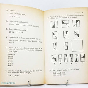 May include: Open book with "TEST SEVEN" printed at the top of the pages. The left page has questions with missing letters, numbers, and words. The right page has geometric shape puzzles. The book is held open by a metal clip.