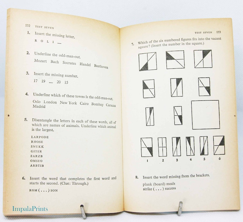 May include: Open book with "TEST SEVEN" printed at the top of the pages. The left page has questions with missing letters, numbers, and words. The right page has geometric shape puzzles. The book is held open by a metal clip.