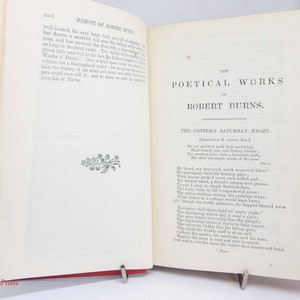 Pode incluir: Um livro antigo aberto intitulado The Poetical Works of Robert Burns, com p&aacute;ginas cor de creme e texto impresso em preto. A p&aacute;gina esquerda mostra uma se&ccedil;&atilde;o de mem&oacute;rias com uma pequena ilustra&ccedil;&atilde;o floral, enquanto a p&aacute;gina direita exibe o poema The Cotter's Saturday Night.