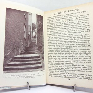 May include: A black and white photograph of an open book featuring a narrow stone staircase leading upwards between brick buildings. The text "CONSTITUTION STEPS, ST. PETER PORT" is visible below the image. The book is held open by a metal clip.