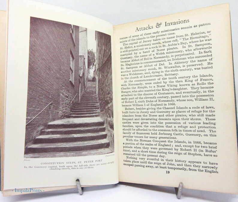 May include: A black and white photograph of an open book featuring a narrow stone staircase leading upwards between brick buildings. The text "CONSTITUTION STEPS, ST. PETER PORT" is visible below the image. The book is held open by a metal clip.