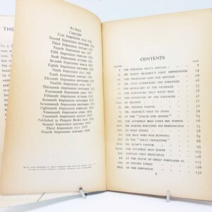 Op de afbeelding: Open boek met inhoudsopgave en copyrightpagina. De vergeelde pagina's tonen hoofdstuktitels zoals 'De aankomst van de vreemdeling' en 'De onzichtbare man verliest zijn humeur'. Het boek wordt opengehouden door een metalen clip.