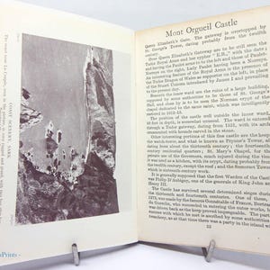 May include: An open book with text and a black and white photograph of a coastal landscape. The text describes Mont Orgueil Castle and coastal scenery on Sark. The book is propped open with a metal book stand.